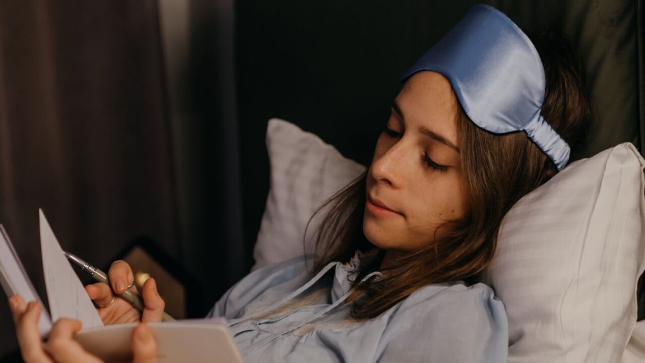A woman in bed with a sleep mask pushed up on her forehead, writing in a notebook